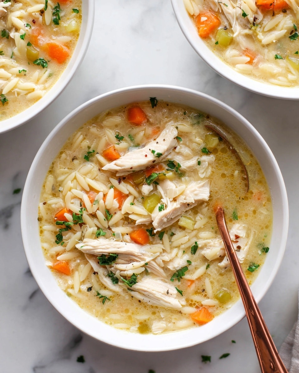 A white bowl filled with creamy chicken soup showing three light-colored chicken pieces on the top layer, scattered orange carrot cubes, and small green herb bits. The soup base is pale yellow and thick, mixed with short, thin white pasta pieces that are visible throughout. A rose-gold spoon rests inside the bowl on the right, with soup, carrots, and pasta on it. A thin lemon slice is placed on the bowl's inner edge to the right. The bowl sits on a white marbled surface, and a second bowl of the same soup is partially visible near the top right corner. photo taken with an iphone --ar 4:5 --v 7